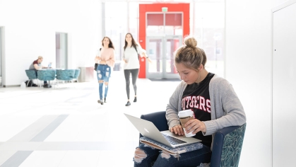 Blonde young female student holding coffee cup and laptop viewing inside