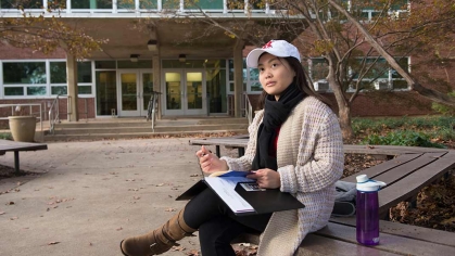 Asian woman with a hat sitting outside, looking up introspectively with folder in hand