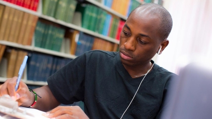 Black student writing in library with earbuds on