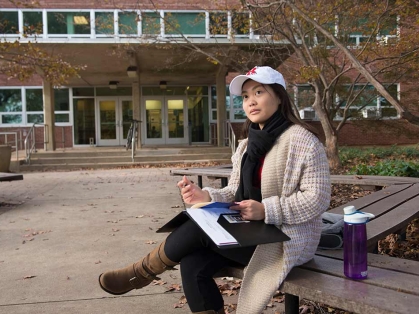 Asian woman with a hat sitting outside, looking up introspectively with folder in hand