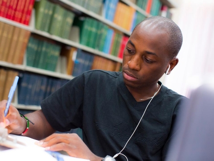 Black student writing in library with earbuds on