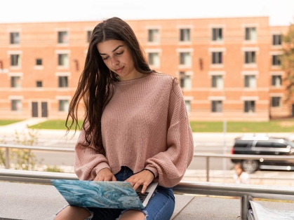female with long hair and pink sweater working on laptop outside